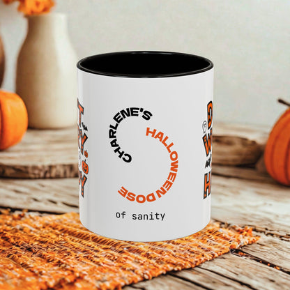 A white ceramic mug with a black interior, printed with the phrase 'Charlene's Halloween Dose of sanity' in black and orange text, styled on a rustic wooden table with autumn props like pumpkins and a woven placemat.