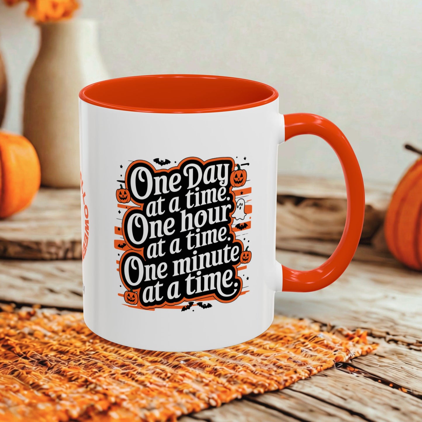 A white ceramic coffee mug with a bright orange handle and interior, featuring a Halloween-themed graphic that reads 'One Day at a time, One hour at a time, One minute at a time.' The mug is styled on a rustic wooden table with autumn props including pumpkins and a woven placemat.