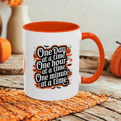 A white ceramic coffee mug with a bright orange handle and interior, featuring a Halloween-themed graphic that reads 'One Day at a time, One hour at a time, One minute at a time.' The mug is styled on a rustic wooden table with autumn props including pumpkins and a woven placemat.