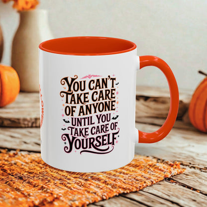 A white ceramic mug with a bright orange handle and interior, featuring a Halloween-themed printed quote in black and pink. The mug is styled on a rustic wooden table with a woven placemat and autumn decorations in the background.