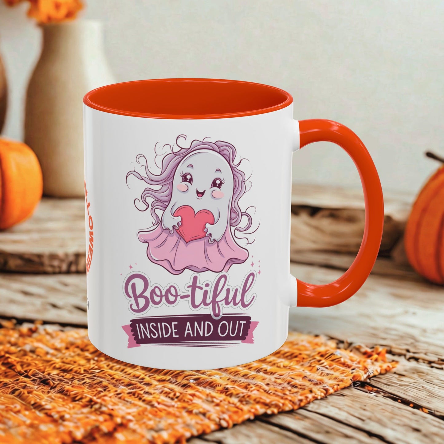 A white ceramic mug with a bright orange handle and interior, featuring a cute cartoon ghost graphic and the text 'Boo-tiful INSIDE AND OUT.' The mug is styled on a woven orange placemat over a rustic wooden surface, with pumpkins and a blurred indoor background.