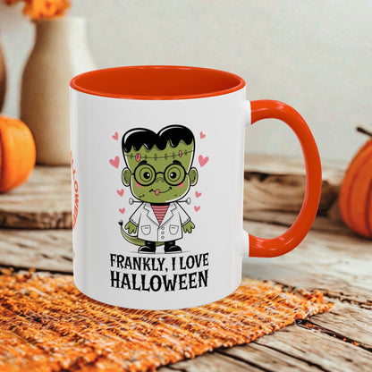 A white ceramic mug with a bright orange interior, handle, and rim, featuring a cartoon Frankenstein character and the text 'FRANKLY, I LOVE HALLOWEEN.' The mug is styled on a woven orange placemat over a rustic wooden surface with pumpkins and a blurred vase in the background.
