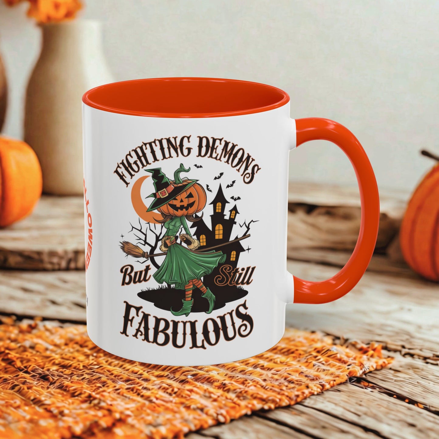 A white ceramic mug with a bright orange handle and interior, featuring a Halloween-themed graphic of a witch with a pumpkin head and the text 'Fighting Demons But Still Fabulous.' The mug is styled on a rustic wooden surface with autumn props in the background.
