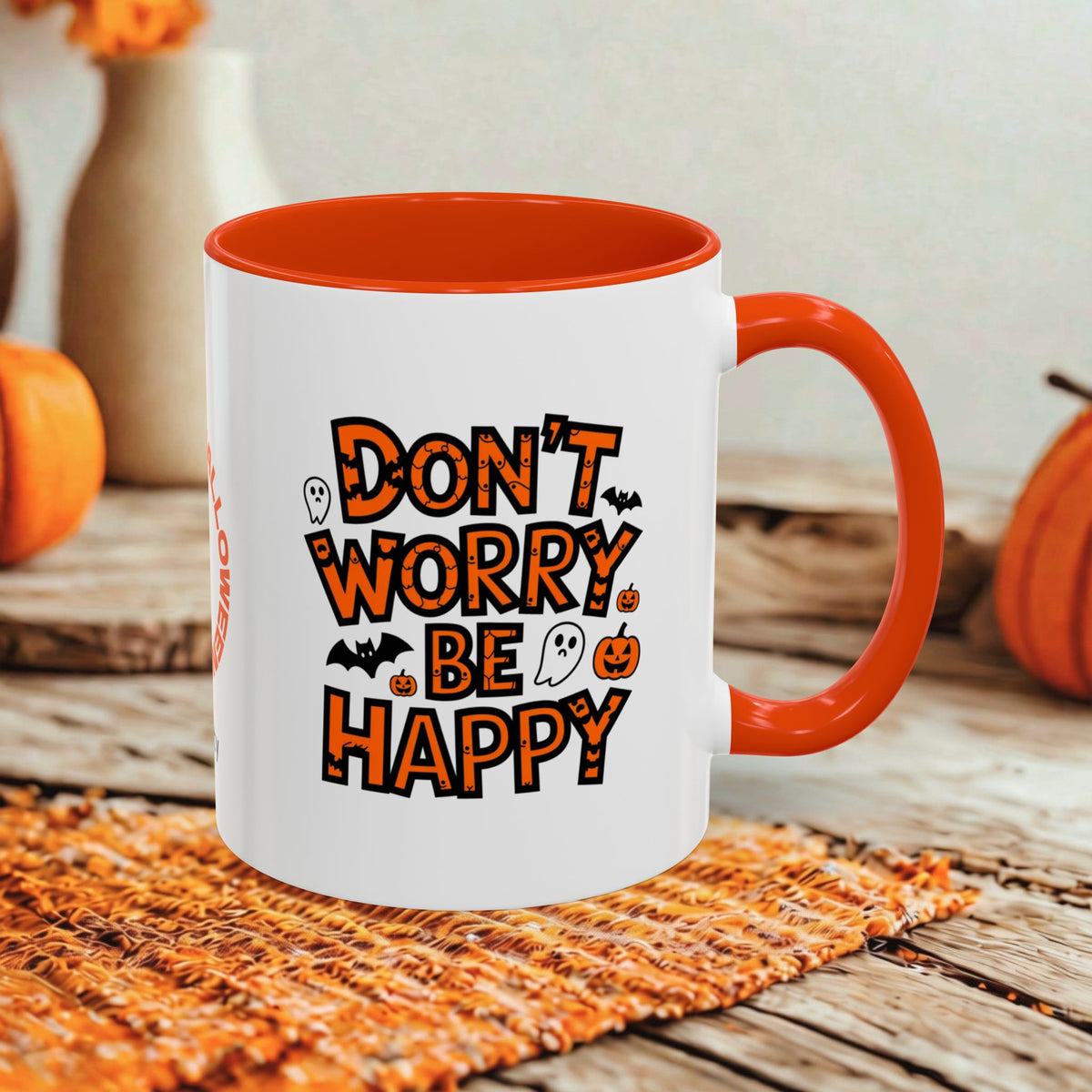 A white ceramic mug with a bright orange handle and interior, featuring the phrase 'DON'T WORRY BE HAPPY' in orange lettering with Halloween-themed illustrations. The mug is set on a rustic wooden table with autumn decorations in the background.