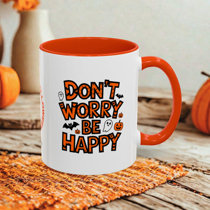 A white ceramic mug with a bright orange handle and interior, featuring the phrase 'DON'T WORRY BE HAPPY' in orange lettering with Halloween-themed illustrations. The mug is set on a rustic wooden table with autumn decorations in the background.
