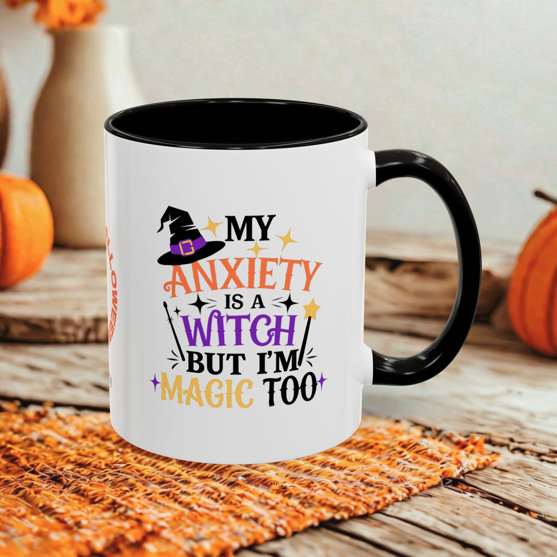 A white ceramic mug with a black handle and interior, printed with the phrase 'MY ANXIETY IS A WITCH BUT I'M MAGIC TOO' in orange, purple, and black lettering with a witch hat graphic. The mug sits on a woven orange placemat on a rustic wooden table, with pumpkins and blurred decorative vases in the background.