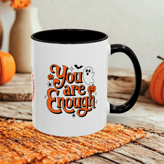A white ceramic mug with a black handle and interior, featuring the phrase 'You are Enough' in orange script with Halloween-themed illustrations including pumpkins, bats, and a ghost. The mug is styled on a woven placemat with autumn pumpkins and a rustic wooden table in the background.