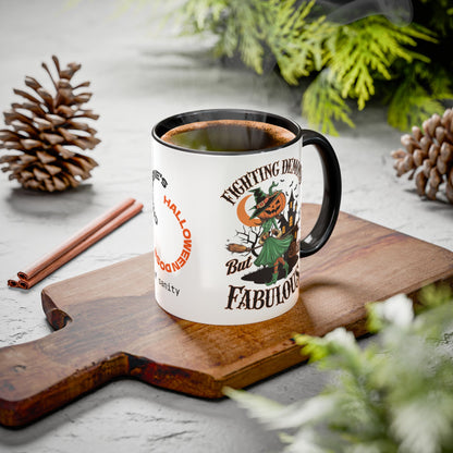 A white ceramic coffee mug with a black handle and rim, featuring a Halloween-themed graphic that reads 'Fighting Demon But Fabulous' with a pumpkin-headed character, styled on a wooden cutting board with pinecones and evergreen branches.