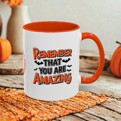 A white ceramic coffee mug with a bright orange handle and interior, featuring the printed text 'REMEMBER THAT YOU ARE AMAZING' with small bat silhouettes. The mug is styled on a rustic wooden surface with a woven orange placemat and blurred pumpkins in the background.