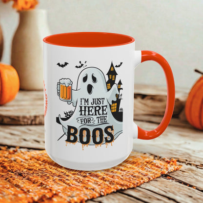 A white ceramic mug with a bright orange handle and interior, featuring a Halloween graphic of a ghost holding a beer mug with the text 'I'M JUST HERE FOR THE BOOS'. The mug is set on a woven orange placemat on a rustic wooden table, with pumpkins and a blurred background.