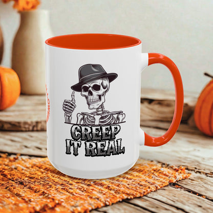 A white ceramic mug with a bright orange handle and interior, featuring a black graphic of a skeleton in a hat giving a thumbs-up with the text 'CREEP IT REAL'. The mug is styled on a rustic wooden surface with autumn-themed props like pumpkins and a woven placemat.