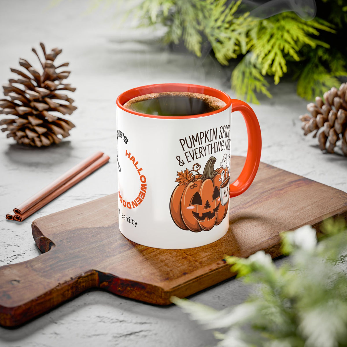 A white ceramic coffee mug with an orange handle and rim, featuring a Halloween-themed graphic with jack-o'-lanterns and the text 'Pumpkin Spice & Everything Nice'. The mug is styled on a wooden cutting board with pinecones, cinnamon sticks, and green foliage on a gray stone surface.