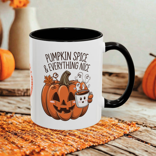 A white ceramic mug with a black handle and interior, printed with a Halloween design featuring a jack-o'-lantern, ghosts, and the text 'PUMPKIN SPICE & EVERYTHING NICE.' The mug is styled on a woven orange placemat over a rustic wooden surface, with blurred pumpkins and a vase in the background.