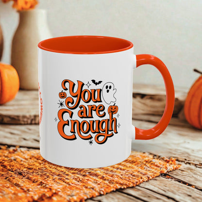 A white ceramic mug with a bright orange interior, handle, and printed graphic reading 'You are Enough' in orange lettering with black outlines, plus small Halloween motifs including pumpkins, bats, stars, and a ghost. The mug is styled on a woven orange mat over a rustic wooden surface with pumpkins and blurred decor in the background.