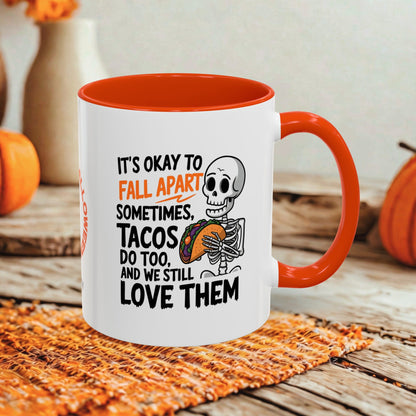 A white ceramic mug with a bright orange interior and handle, featuring a Halloween-themed printed design with a skeleton holding a taco and the text 'IT'S OKAY TO FALL APART SOMETIMES. TACOS DO TOO.