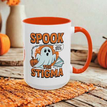 A white ceramic mug with a bright orange handle and interior, featuring a Halloween graphic that reads 'SPOOK THE STIGMA' with a ghost and pumpkin illustration. The mug is styled on a rustic wooden surface with autumn props in the background.