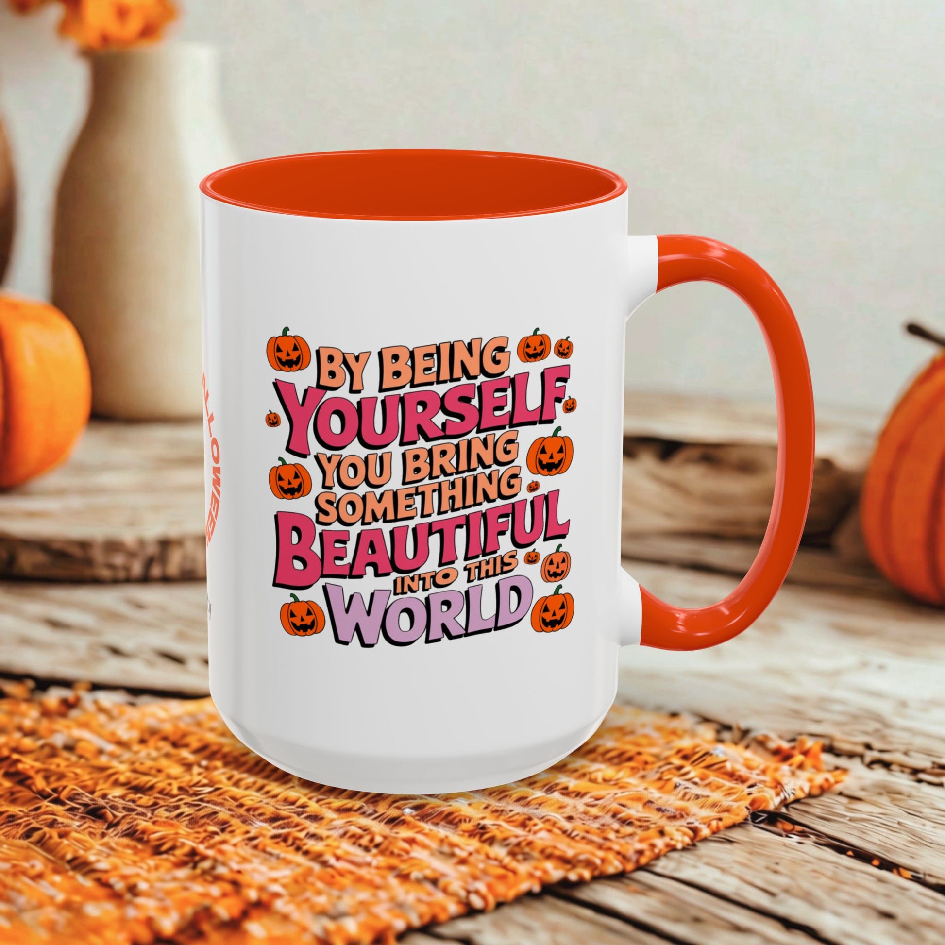 A white ceramic mug with an orange interior and handle, featuring a colorful Halloween-themed quote that reads 'BY BEING YOURSELF YOU BRING SOMETHING BEAUTIFUL INTO THE WORLD' surrounded by jack-o'-lantern illustrations. The mug is styled on a rustic wooden table with autumn props including pumpkins and a woven placemat.