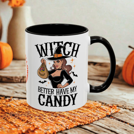 A white ceramic mug with a black handle and interior, printed with the phrase 'WITCH BETTER HAVE MY CANDY' and an illustration of a witch holding a bag of candy. The mug is styled on a rustic wooden surface with autumn props including pumpkins and a woven placemat.