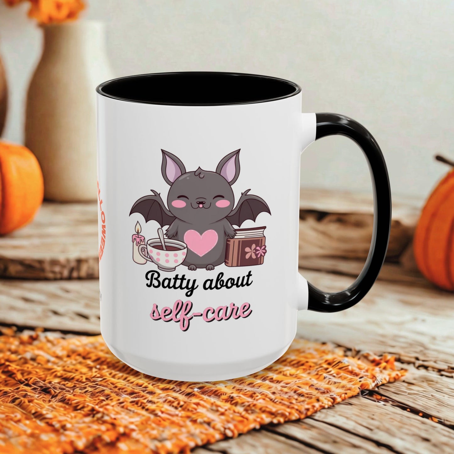 A white ceramic mug with a black handle and black interior, printed with a cute bat graphic and the phrase 'Batty about self-care' in black and pink lettering. The mug is styled on a rustic wooden surface with autumn-themed props like pumpkins and a woven placemat.