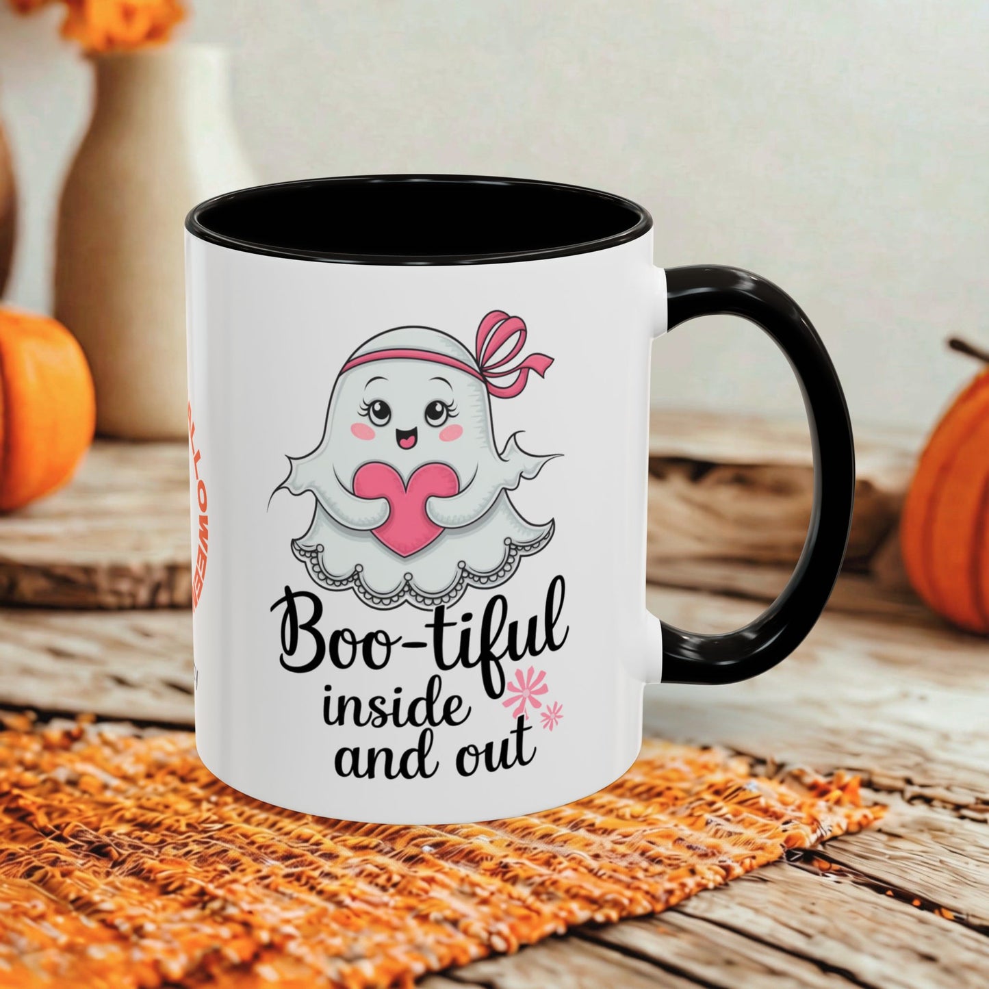 A white ceramic mug with a black handle and black interior, printed with a cute ghost holding a pink heart and the phrase 'Boo-tiful inside and out.' The mug is styled on a woven orange placemat over a rustic wooden surface, with pumpkins and a blurred indoor background.