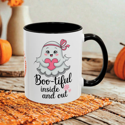 A white ceramic mug with a black handle and black interior, printed with a cute ghost holding a pink heart and the phrase 'Boo-tiful inside and out.' The mug is styled on a woven orange placemat over a rustic wooden surface, with pumpkins and a blurred indoor background.