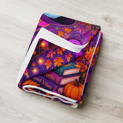 A colorful Halloween-themed blanket with the phrase “Treat Yourself Kindly” in large decorative lettering. The scene shows a smiling ghost in a witch hat holding a mug, surrounded by pumpkins, flowers, a house, candles, and autumn leaves on a purple background.