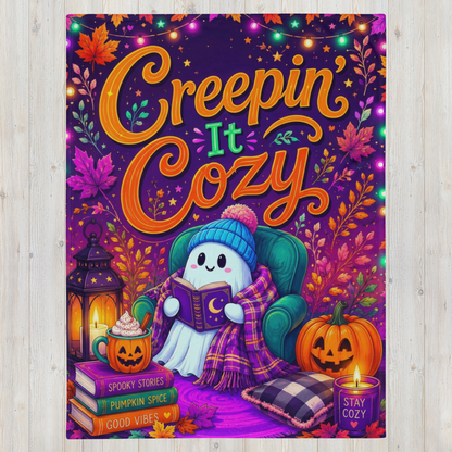 A colorful Halloween-themed blanket featuring the phrase 'Creepin' It Cozy' in large orange lettering. The illustration shows a cute white ghost character wearing a blue knit hat and plaid blanket, sitting in a green armchair and reading a book.
