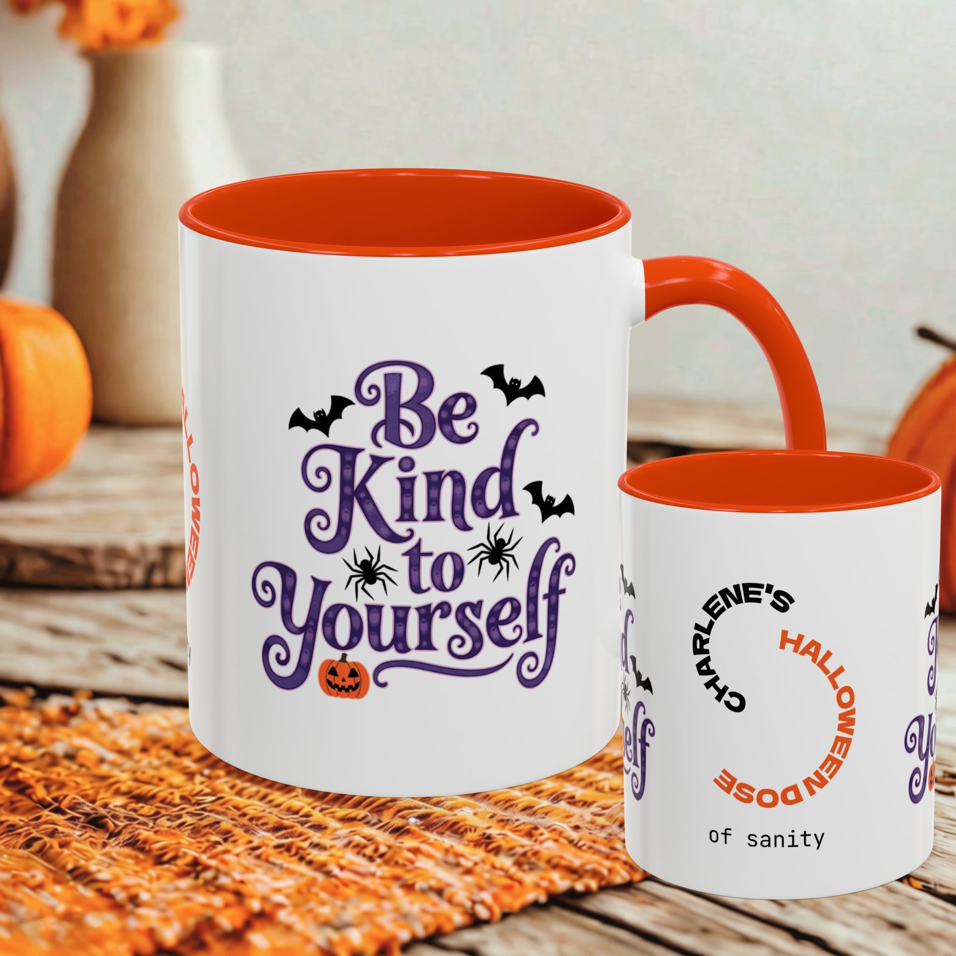 Two white ceramic mugs with orange interiors and handles, styled on a woven orange placemat with autumn decor in the background. The larger mug reads 'Be Kind to Yourself' in purple script with black bats and a small orange jack-o'-lantern.