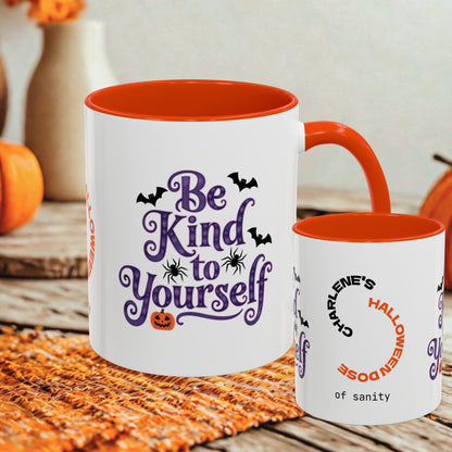 Two white ceramic mugs with orange interiors and handles, styled on a woven orange placemat with autumn decor in the background. The larger mug reads 'Be Kind to Yourself' in purple script with black bats and a small orange jack-o'-lantern.