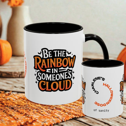 Two white ceramic mugs with black interiors and handles, displayed on a rustic wooden surface with autumn props. The larger mug shows the text 'Be the Rainbow in Someone's Cloud' in orange and black lettering, and the smaller mug shows partial text including 'Charlene's Halloween of sanity'.