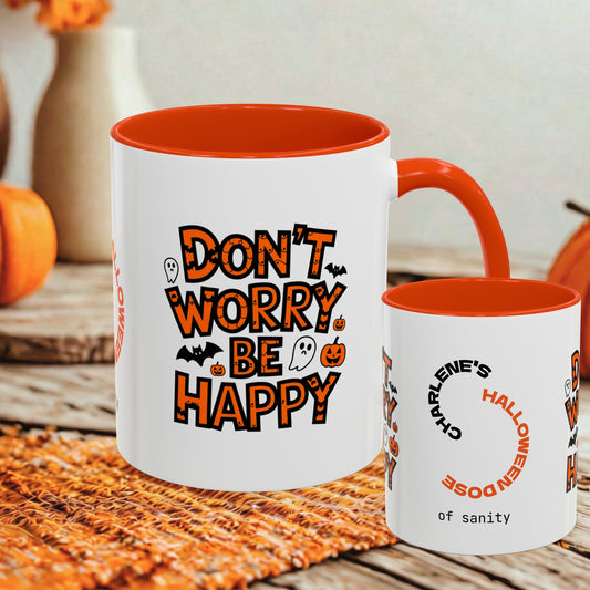 Two white ceramic mugs with orange interiors and handles, styled on a woven orange mat with autumn props in the background. The larger mug reads “DON’T WORRY BE HAPPY” in orange lettering with Halloween graphics, and the smaller mug reads “CHARLIE’S HALLOWEENDOSE of sanity.