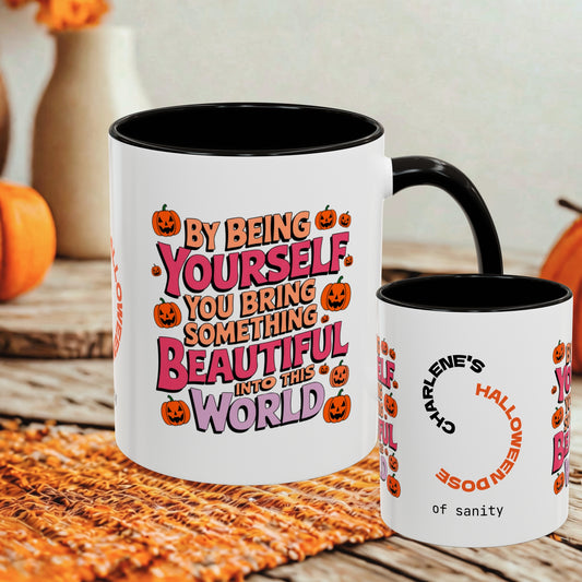 Two white ceramic mugs with black interiors and handles, displayed on a woven orange mat with autumn decor in the background. The larger mug features a colorful Halloween-themed quote, and the smaller mug shows a custom name print.
