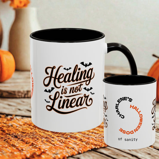 Two white ceramic mugs with black interiors and handles are shown on a woven orange placemat. The larger mug reads “Healing is not Linear” in dark brown script with small bat silhouettes, and the smaller mug has Halloween-themed text including “Charlene’s Halloween dose of sanity.