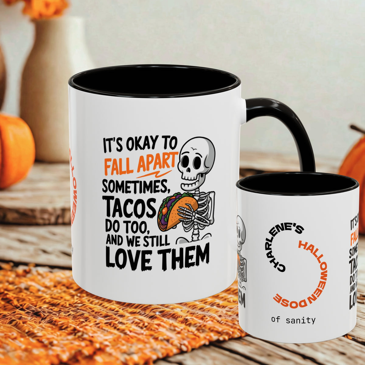 Two white ceramic mugs with black interiors and handles are shown on a woven orange mat in a fall-themed setting. The larger mug features a skeleton holding a taco and the text, 'IT'S OKAY TO FALL APART SOMETIMES.