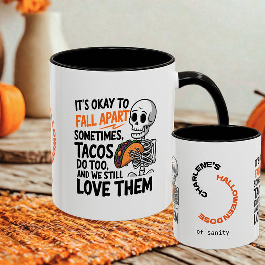 Two white ceramic mugs with black interiors and handles are shown on a woven orange mat in a fall-themed setting. The larger mug features a skeleton holding a taco and the text, 'IT'S OKAY TO FALL APART SOMETIMES.