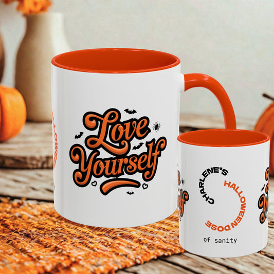 Two white ceramic mugs with orange interiors and handles, styled on a woven placemat with autumn props. The larger mug reads 'Love Yourself' in orange script with small bat and spider graphics, and the smaller mug reads 'Charlene's Halloween of sanity' in orange and black text.