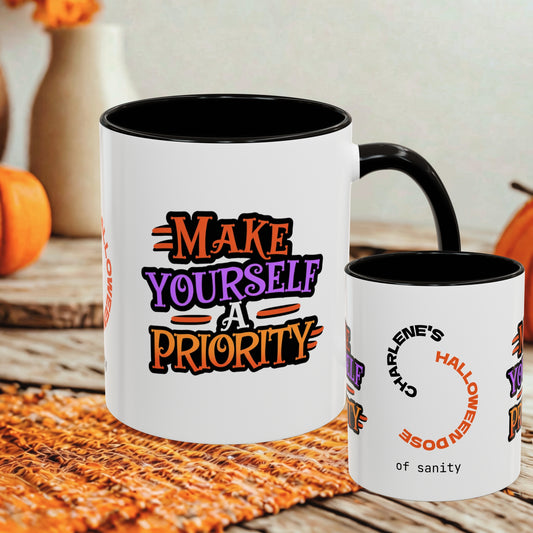 Two white ceramic mugs with black interiors and handles, displayed on a rustic wooden surface with autumn props. The larger mug shows the text 'MAKE YOURSELF A PRIORITY' in orange, purple, and black lettering.