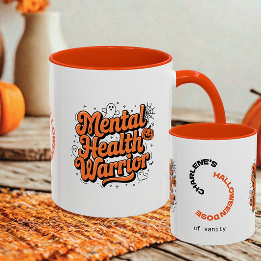 A white ceramic mug with an orange interior and handle, shown alongside a smaller matching cup. The larger mug features the phrase 'Mental Health Warrior' in orange lettering with Halloween-themed doodles, and the smaller cup reads 'Charlene's Halloween Doze of sanity'.