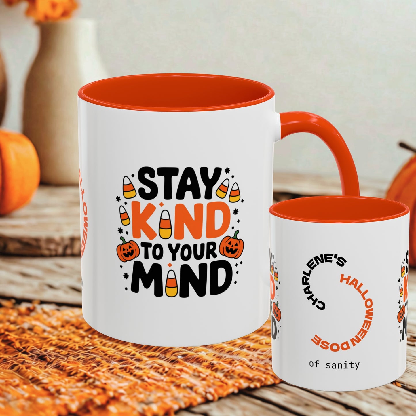 Two white ceramic mugs with orange interiors and handles, styled on a woven placemat with autumn props. The larger mug reads “STAY KIND TO YOUR MIND” with candy corn and pumpkin graphics, and the smaller mug has a Halloween-themed printed design.