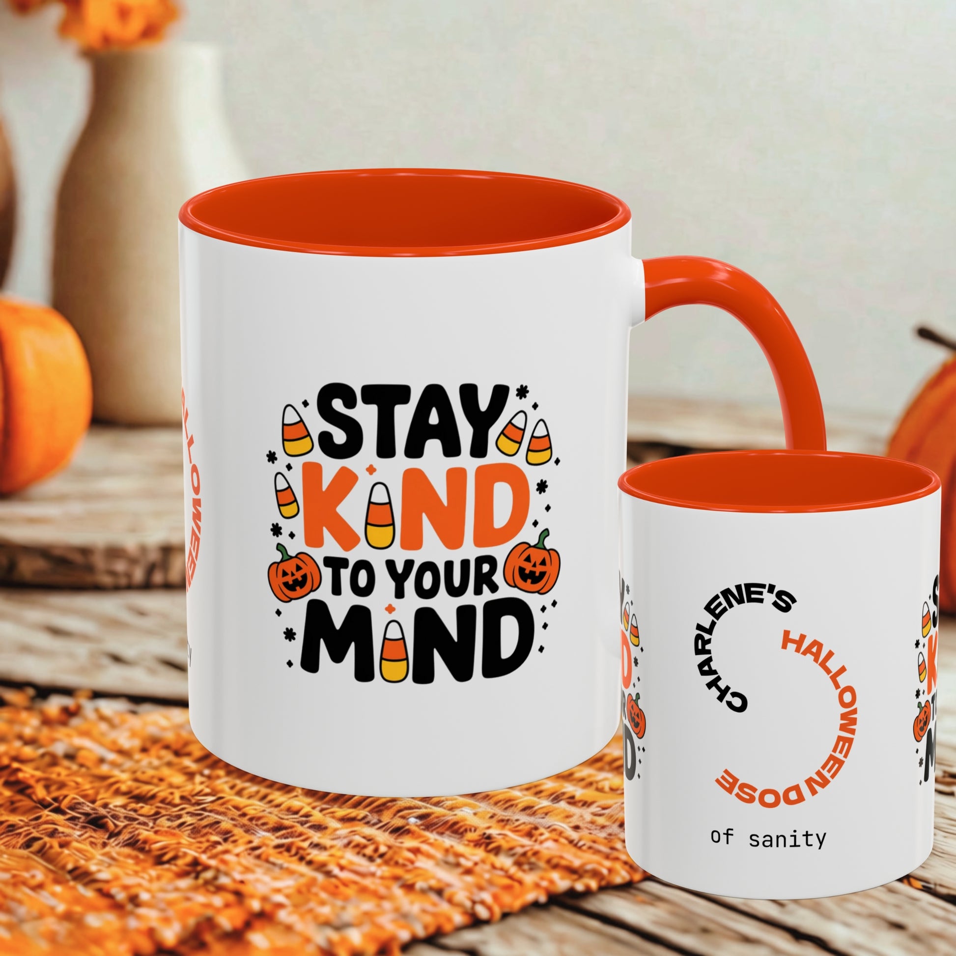 Two white ceramic mugs with orange interiors and handles, styled on a woven placemat with autumn props. The larger mug reads “STAY KIND TO YOUR MIND” with candy corn and pumpkin graphics, and the smaller mug has a Halloween-themed printed design.