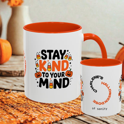 Two white ceramic mugs with orange interiors and handles, styled on a woven placemat with autumn props. The larger mug reads “STAY KIND TO YOUR MIND” with candy corn and pumpkin graphics, and the smaller mug has a Halloween-themed printed design.