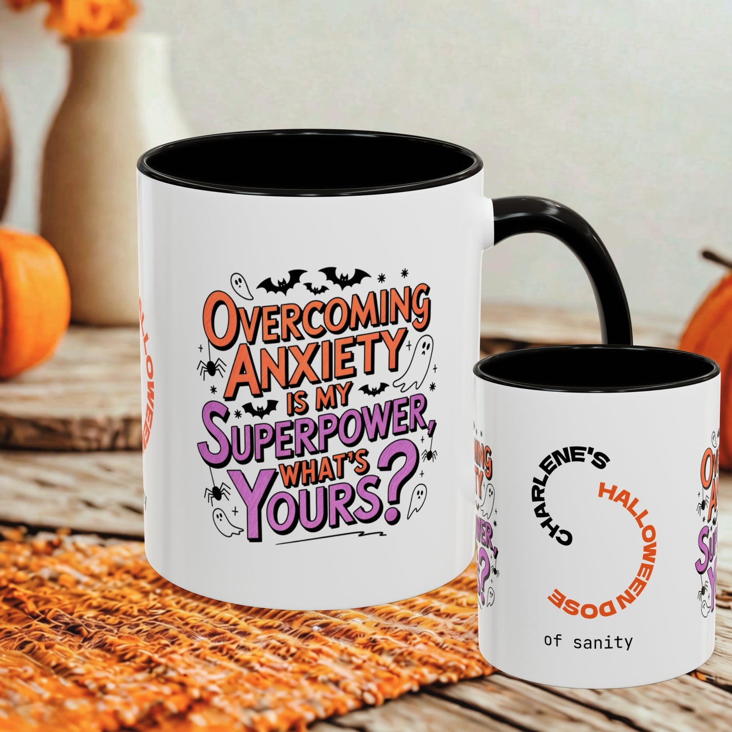 Two white ceramic mugs with black interiors and handles are shown on a rustic table with autumn decor in the background. The larger mug features the phrase 'Overcoming Anxiety Is My Superpower, What's Yours?' in orange, purple, and black lettering, while the smaller mug reads 'Charlene's Halloween Doze of sanity' with a circular arrow graphic.
