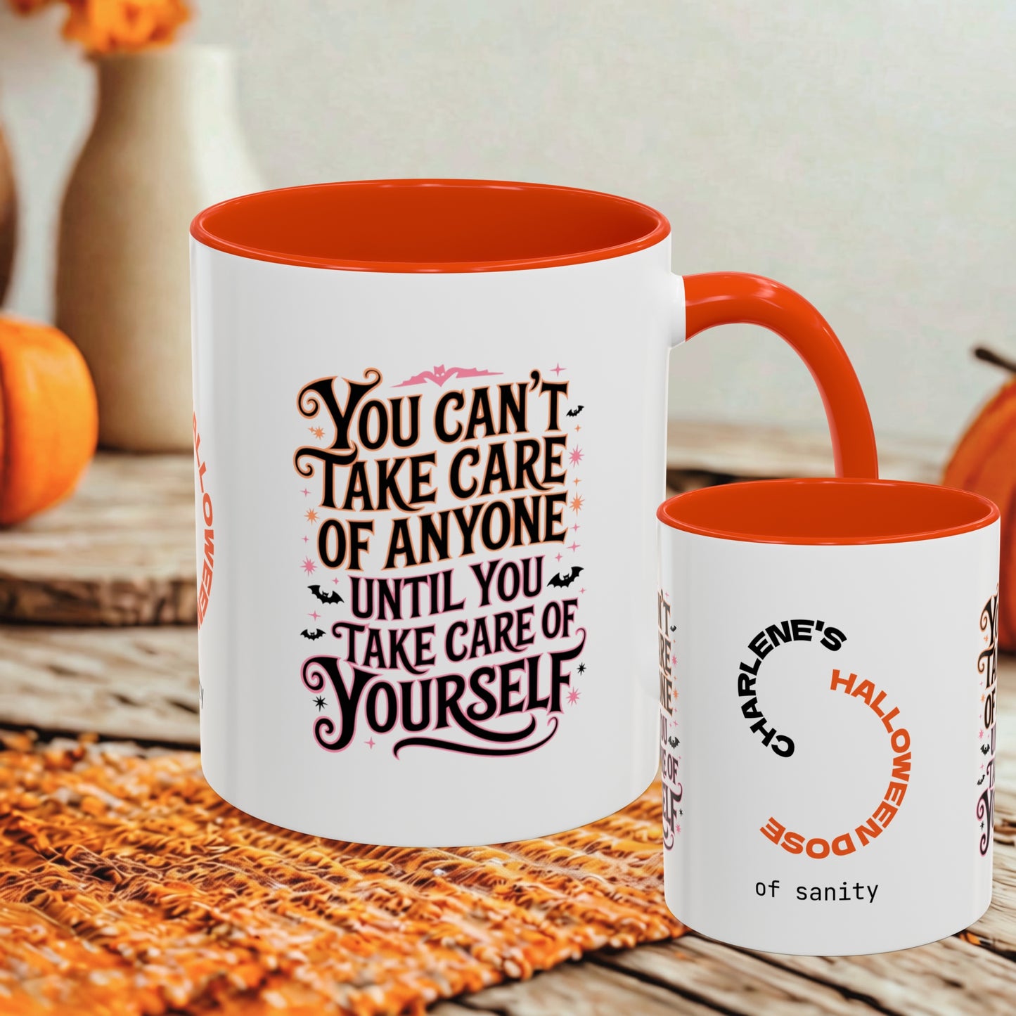 A white ceramic mug with a bright orange interior and handle is shown on a woven orange placemat, with a smaller matching cup beside it. The mugs have Halloween-themed printed text in orange, black, and pink.