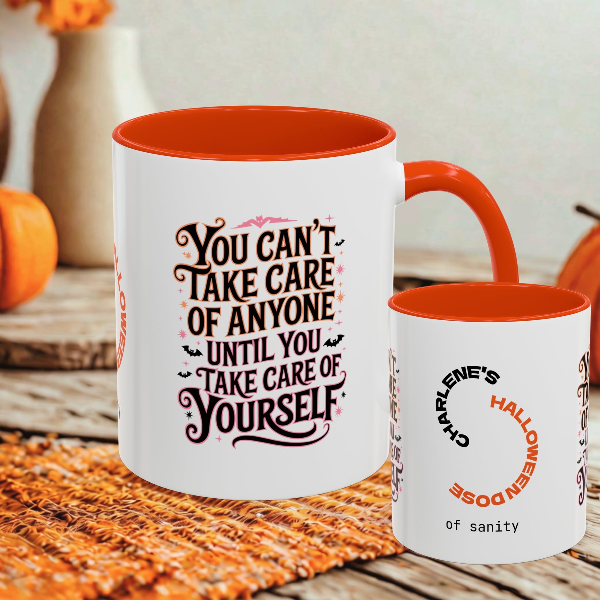 A white ceramic mug with a bright orange interior and handle is shown on a woven orange placemat, with a smaller matching cup beside it. The mugs have Halloween-themed printed text in orange, black, and pink.
