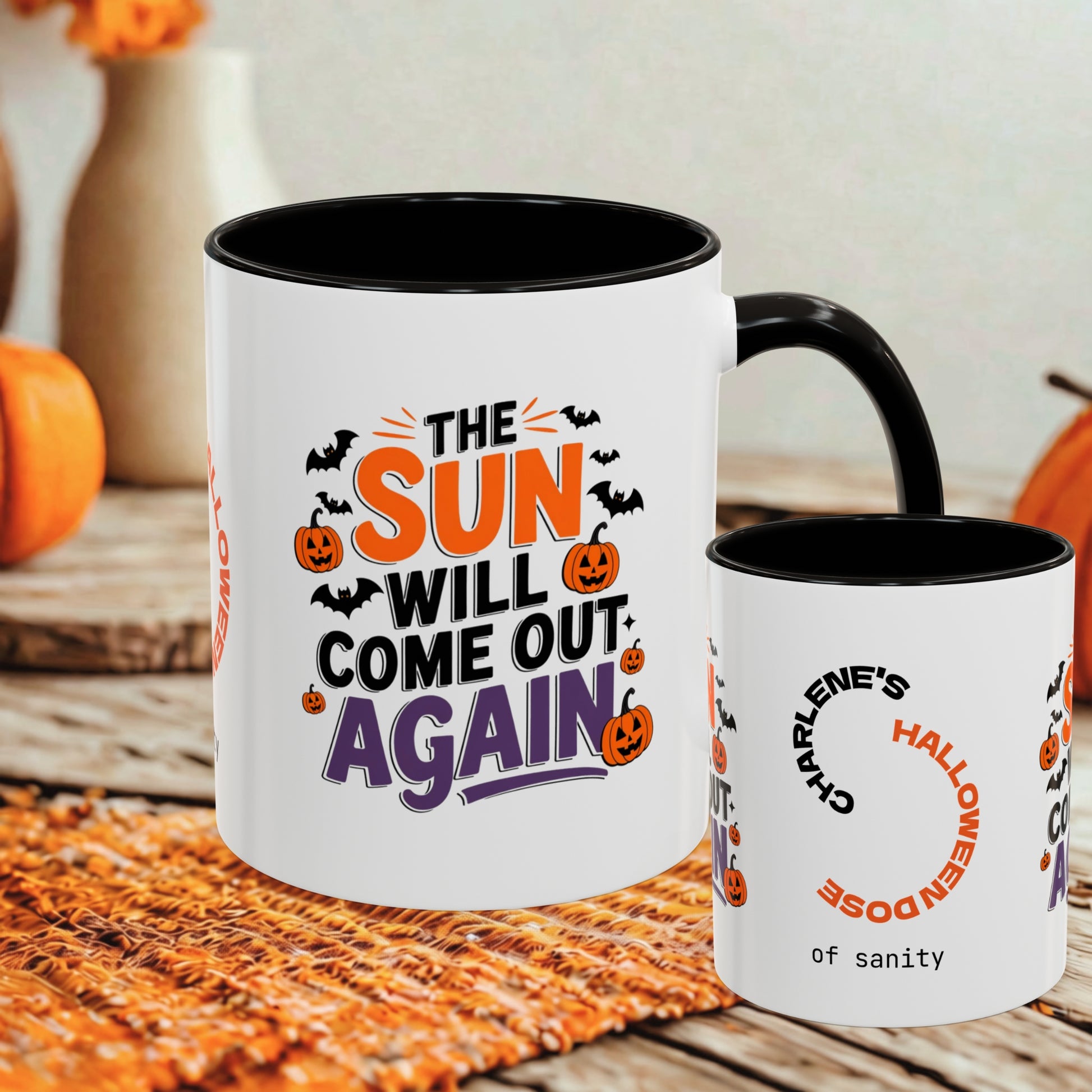 Two white ceramic mugs with black interiors and handles, displayed on a woven orange mat with autumn decor in the background. The larger mug features the phrase “THE SUN WILL COME OUT AGAIN” in orange, purple, and black, with Halloween graphics.