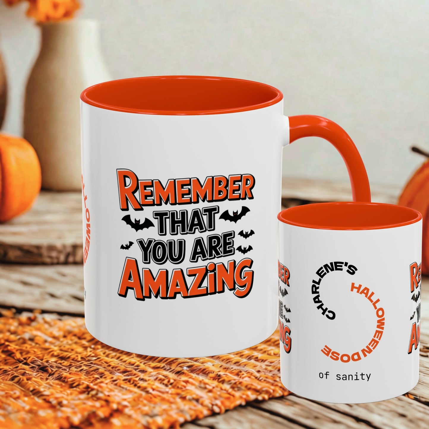 Two white ceramic mugs with orange interiors and handles, displayed on a woven orange placemat with pumpkins and a blurred indoor background. The larger mug shows the text 'REMEMBER THAT YOU ARE AMAZING' with bats, and the smaller mug shows partial text including 'CHARLIE'S HALLOWEENDOSE of sanity'.