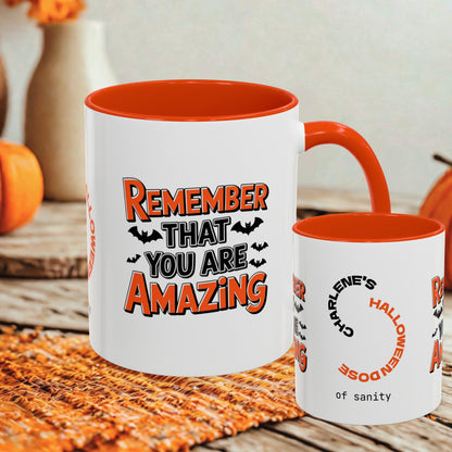 Two white ceramic mugs with orange interiors and handles, displayed on a woven orange placemat with pumpkins and a blurred indoor background. The larger mug shows the text 'REMEMBER THAT YOU ARE AMAZING' with bats, and the smaller mug shows partial text including 'CHARLIE'S HALLOWEENDOSE of sanity'.