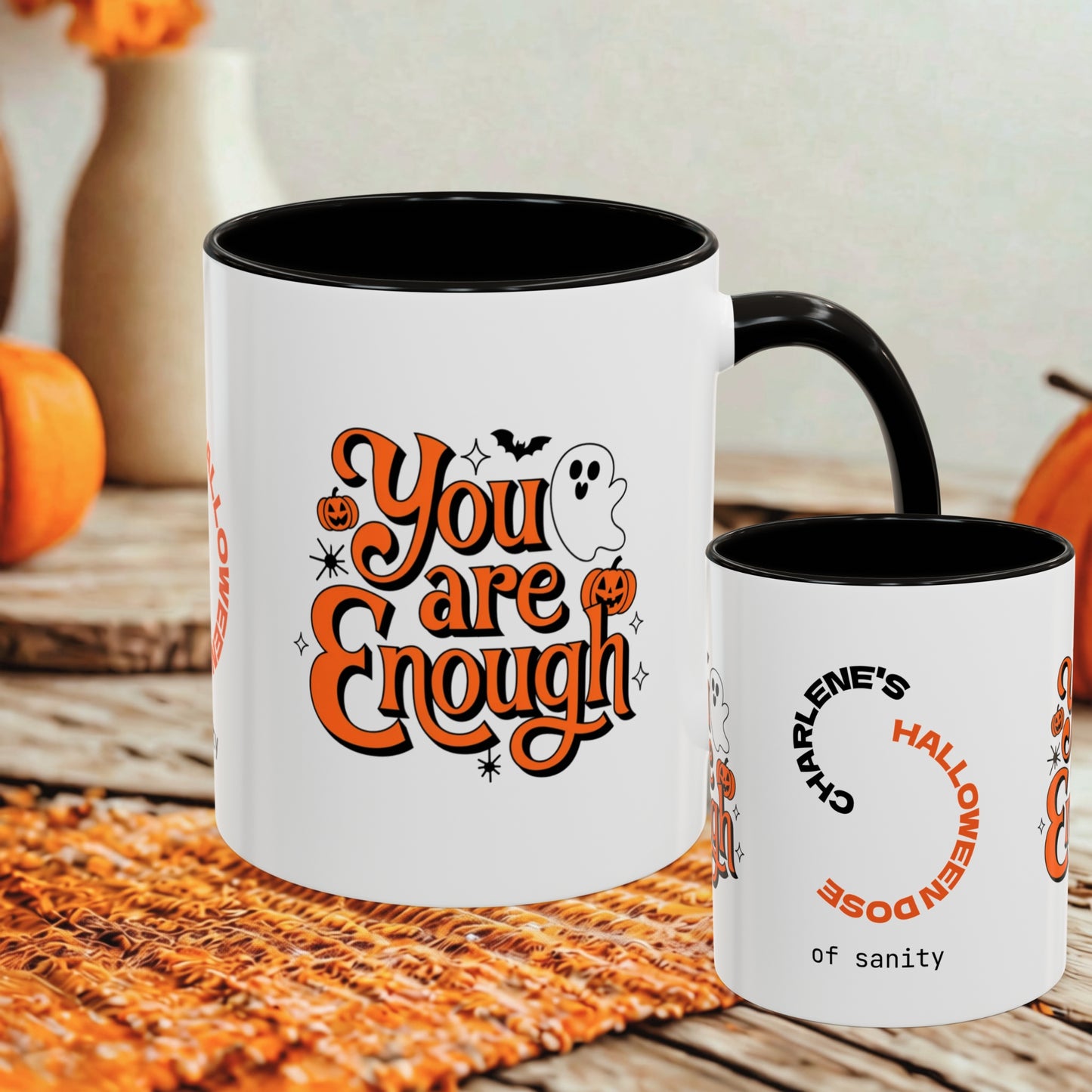 A white ceramic mug with a black interior and handle is shown on a rustic wooden surface with autumn props. The front of the mug features the phrase 'You are Enough' in orange lettering with a ghost, pumpkins, and bats graphic.