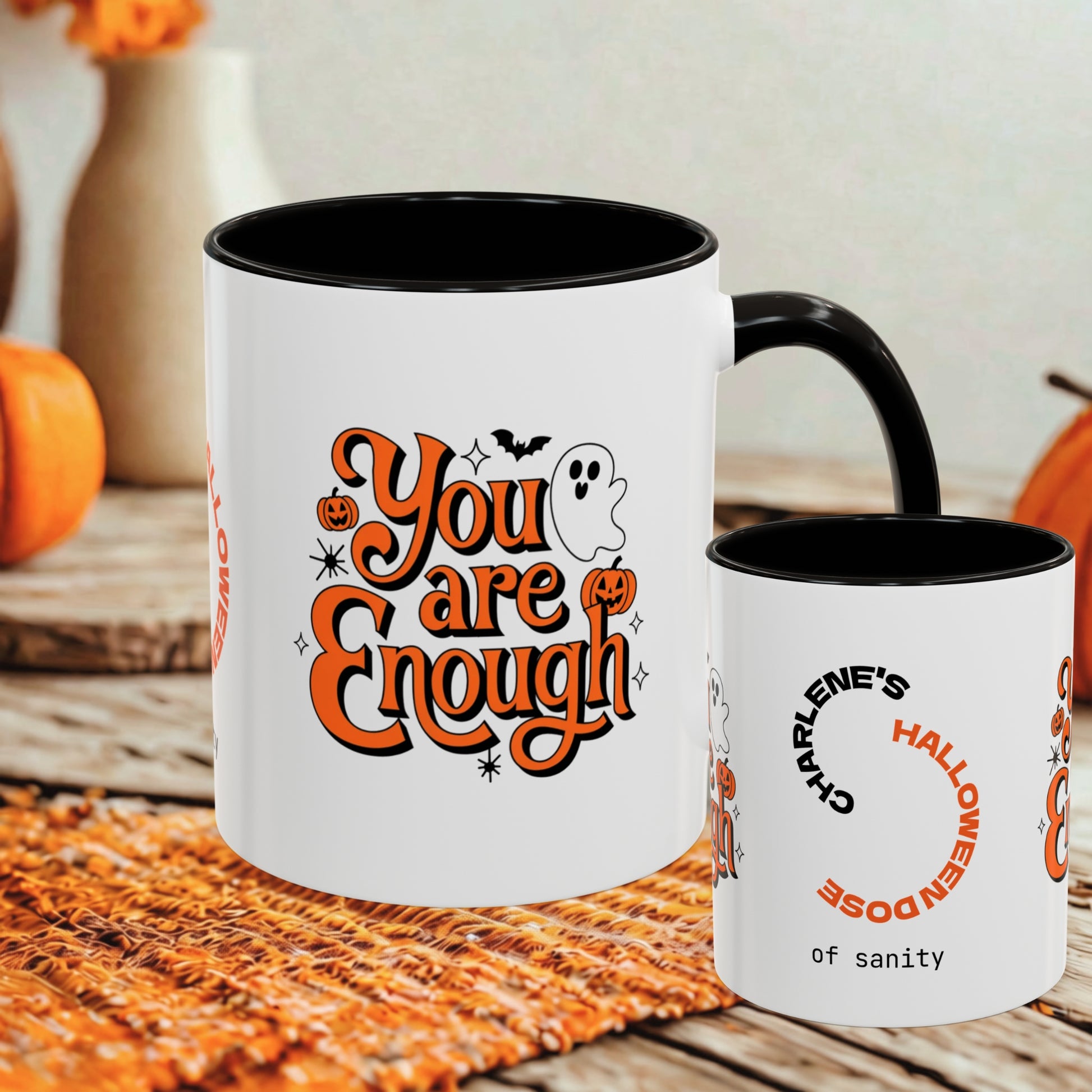 A white ceramic mug with a black interior and handle is shown on a rustic wooden surface with autumn props. The front of the mug features the phrase 'You are Enough' in orange lettering with a ghost, pumpkins, and bats graphic.
