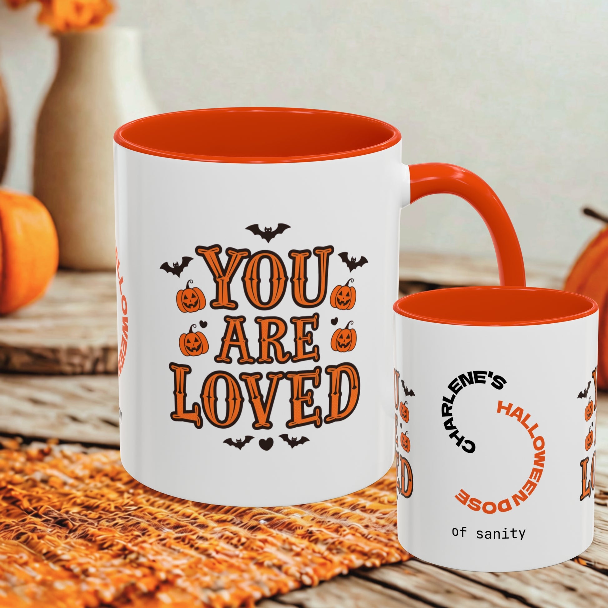 A white ceramic mug with an orange interior and handle, printed with the phrase 'YOU ARE LOVED' in orange lettering with Halloween-themed graphics, shown alongside a smaller matching cup on a rustic wooden surface.